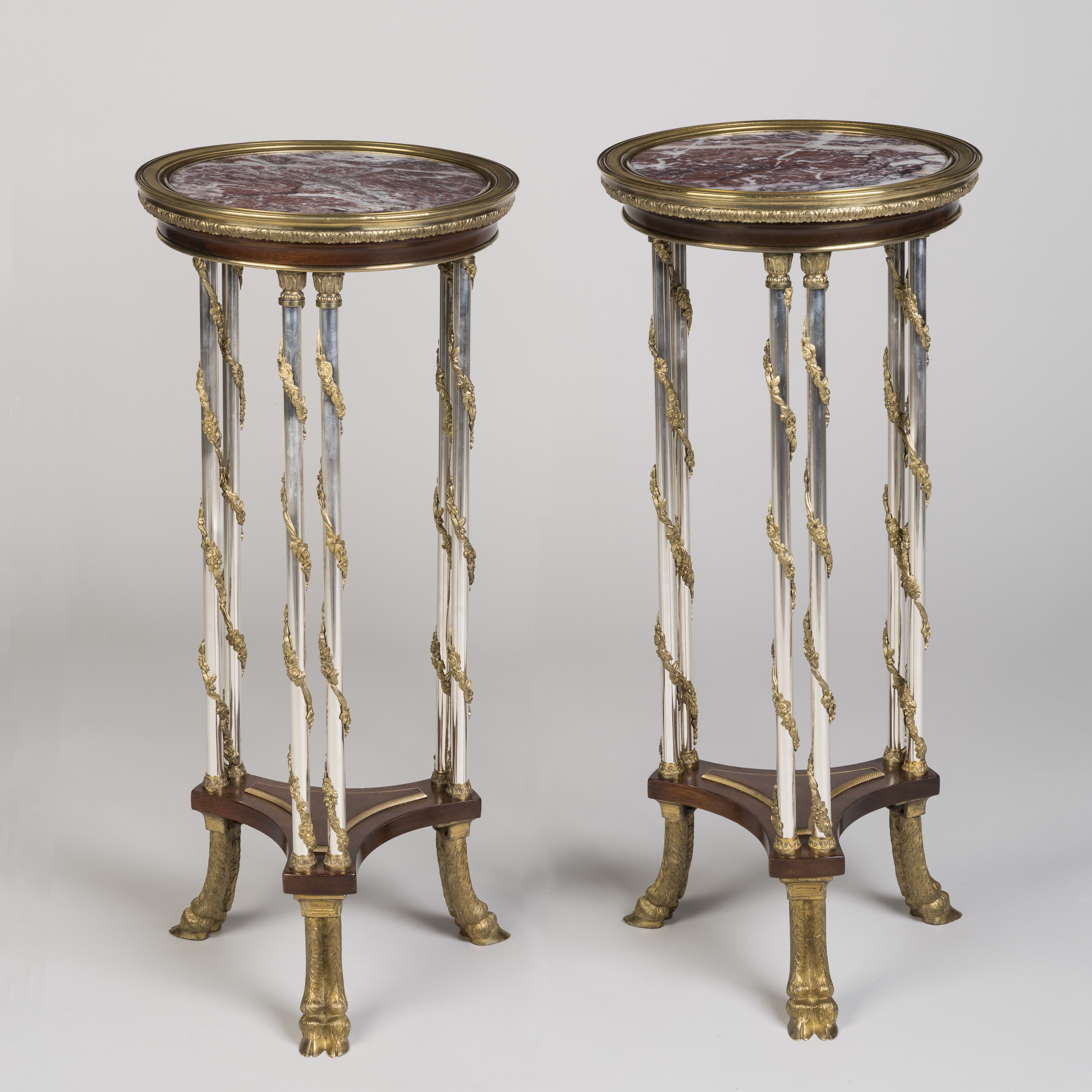 Alt Text: A pair of tall, ornate antique French gueridons (pedestal tables) featuring red and white marbled tops, clear glass supports with gilded floral garlands, and gilt-bronze animal paw feet, set against a plain light grey background.