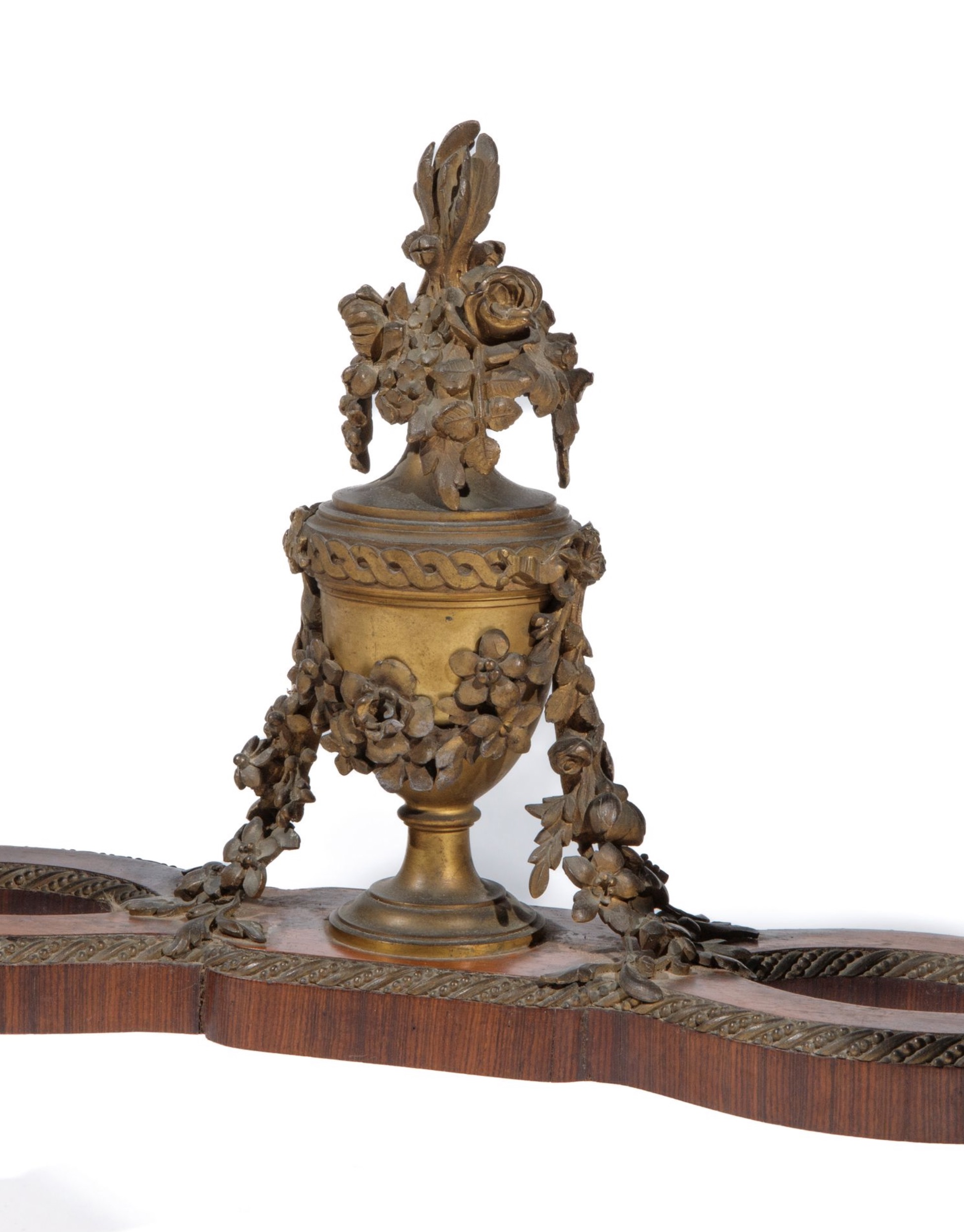 An ornamental bronze urn with draped floral garlands and a rose-topped lid sits centered on a mahogany shelf, featuring intricate casting and a weathered patina against a white background.