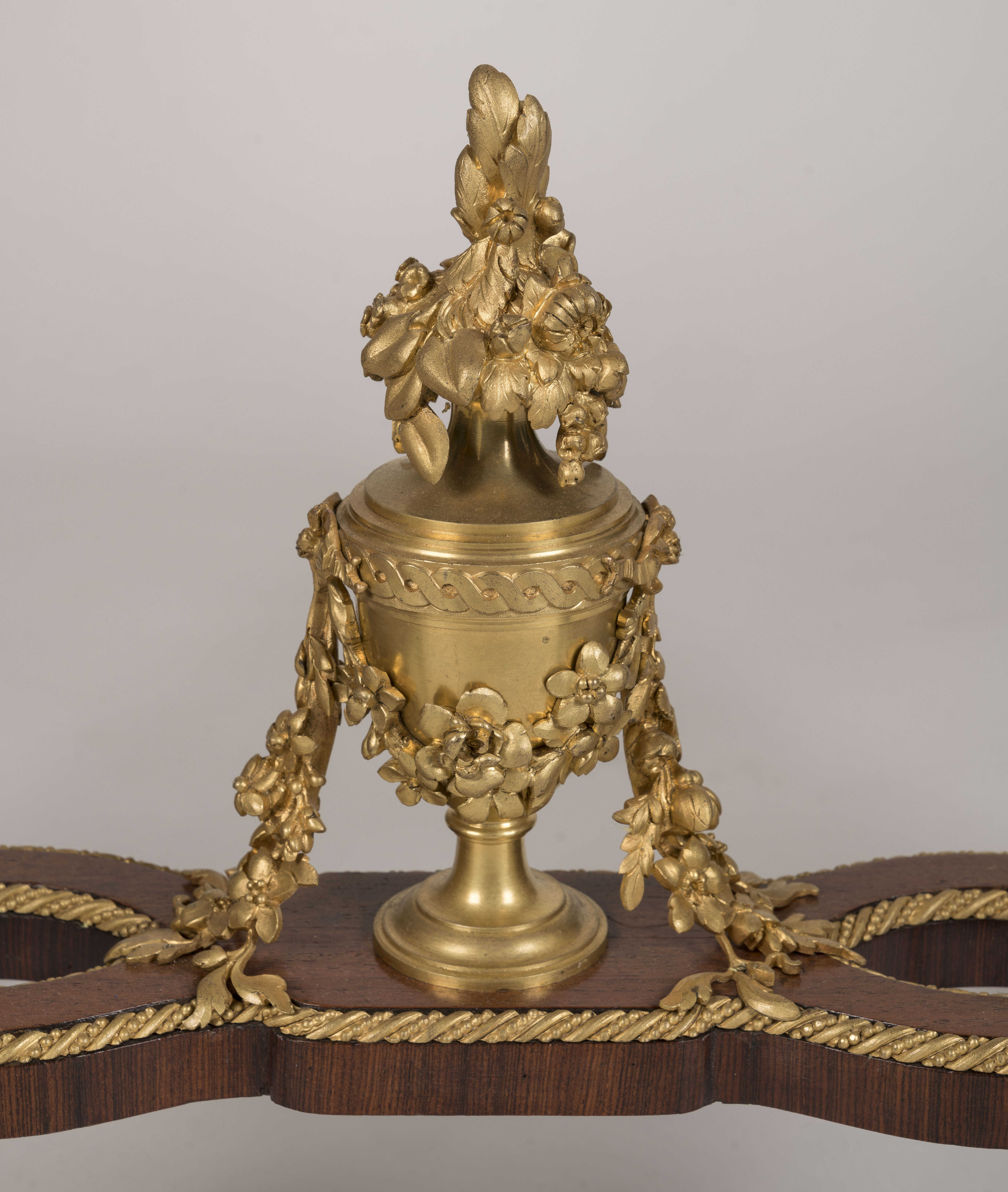 A gilded metal urn with elaborate floral swags and a blossoming finial sits centered on a mahogany stretcher, flanked by twisted rope-style gold molding against a neutral background.