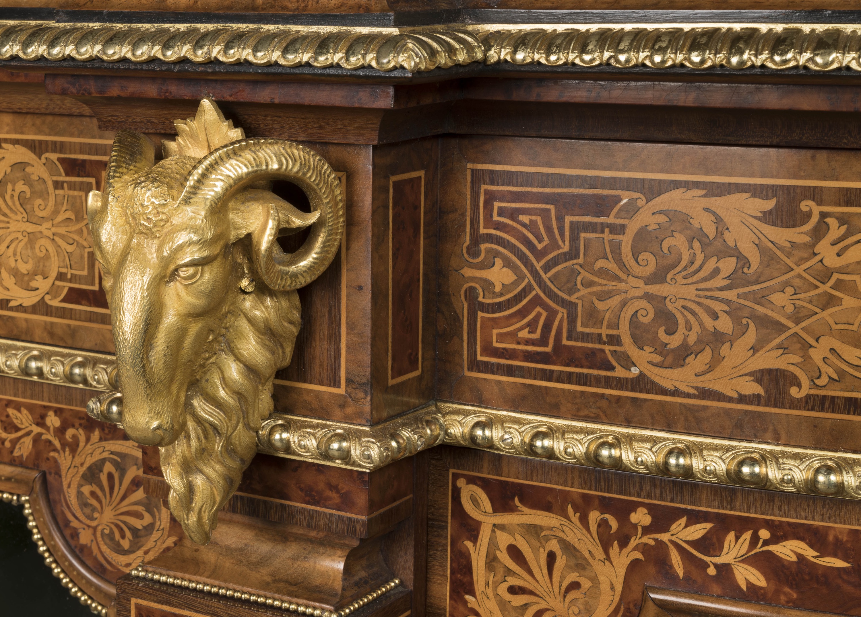 A gilded ram's head mount projects from an inlaid wooden cabinet corner, bordered by gold beading and intricate marquetry scrollwork, showcasing detailed craftsmanship against a dark, polished furniture surface.