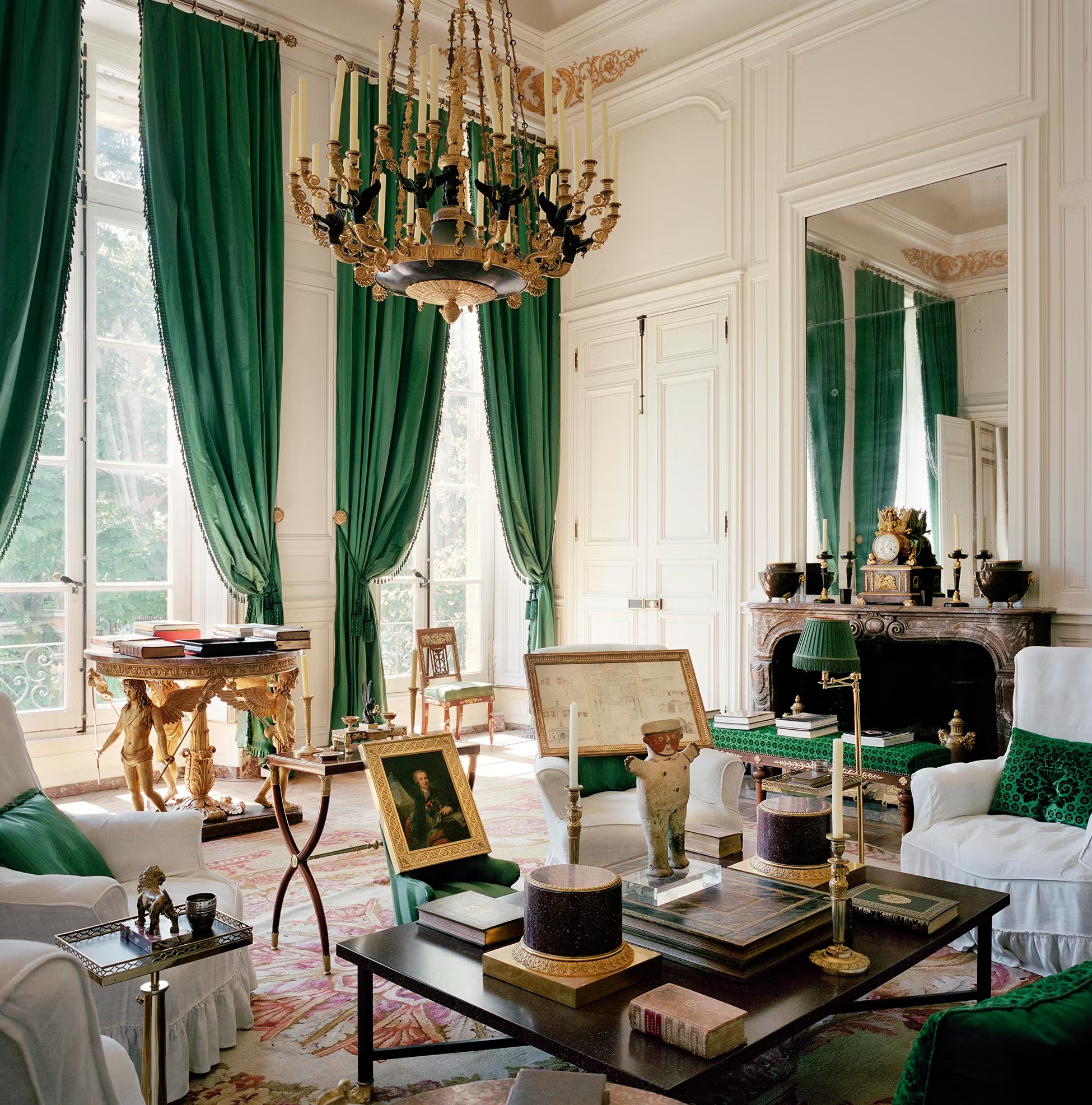 A lavishly decorated Neoclassical salon featuring high white paneled walls, floor-to-ceiling emerald green silk curtains, and a large golden chandelier. The room is filled with a curated mix of Empire-style furniture, including white slipcovered armchairs, a marble fireplace with a gilded clock, and a variety of art objects and books arranged on a central coffee table.