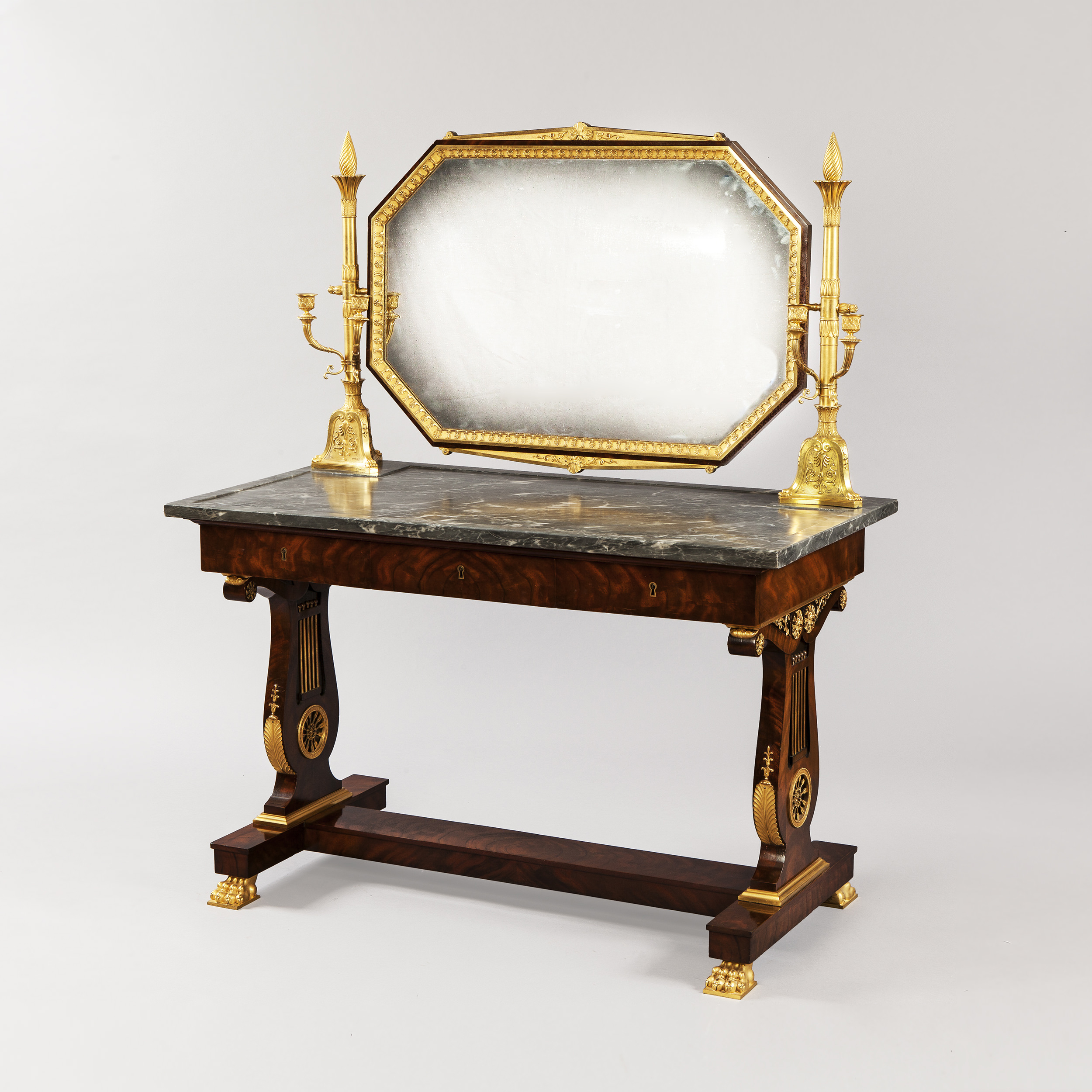 Alt Text (Concise) A French Empire mahogany dressing table attributed to Jacob-Desmalter, featuring a dark grey marble top and a large, octagonal swivel mirror framed in gilded wood. The table is supported by two lyre-shaped pedestal legs adorned with ornate ormolu mounts, connected by a low stretcher and resting on gilded lion-paw feet. Two tall, gilded bronze candelabra stand on the marble surface flanking the mirror.