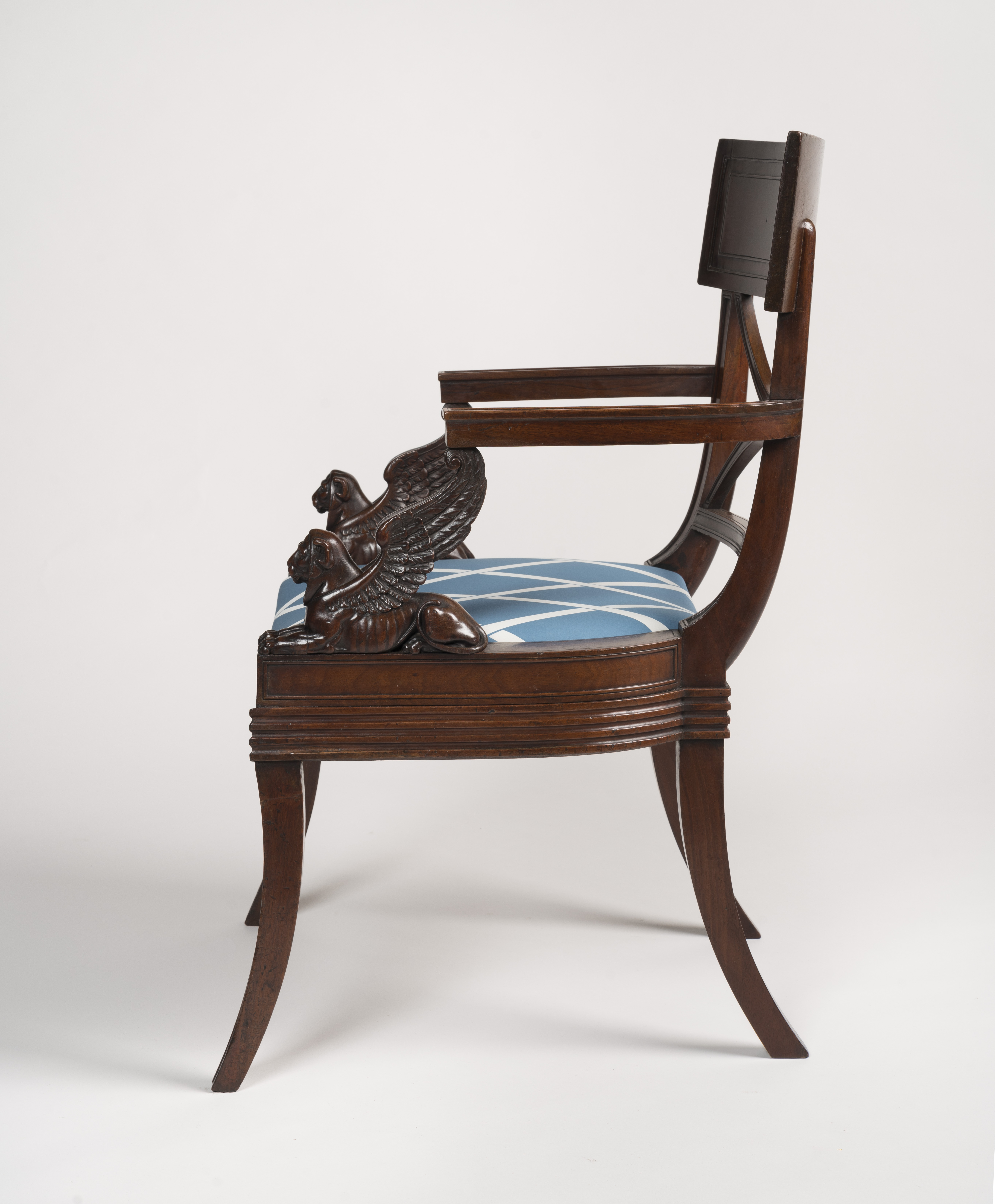 A side-profile view of a Regency-style mahogany armchair, designed after Thomas Hope. The chair features a curved backrest, saber legs, and a distinctive arm support carved in the shape of a winged sphinx. The seat is upholstered in a light blue fabric with a white diamond trellis pattern.