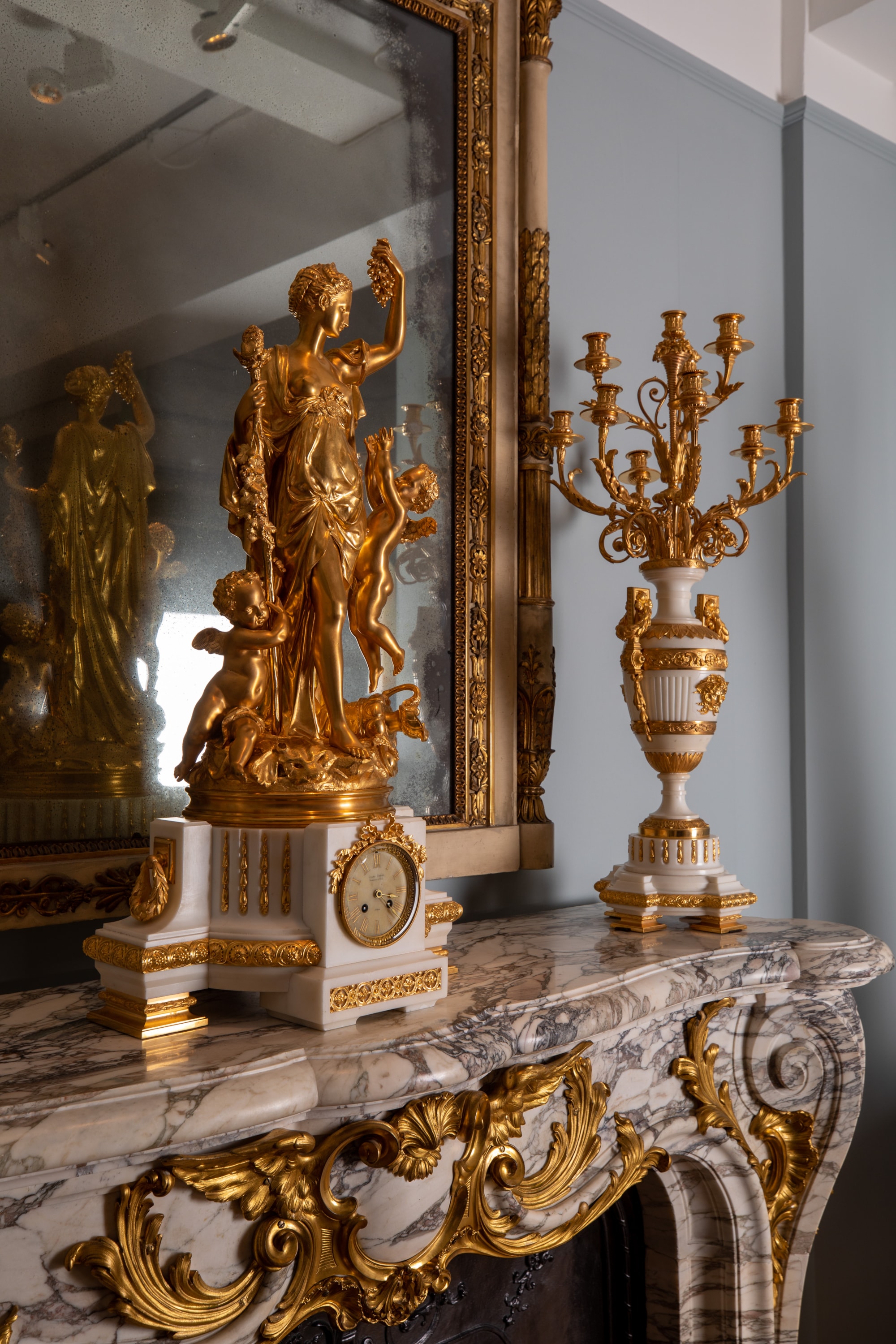 A gilded neoclassical clock featuring a female figure and cherubs stands next to a white marble candelabrum on a marble mantelpiece, positioned before a large gold-framed mirror.