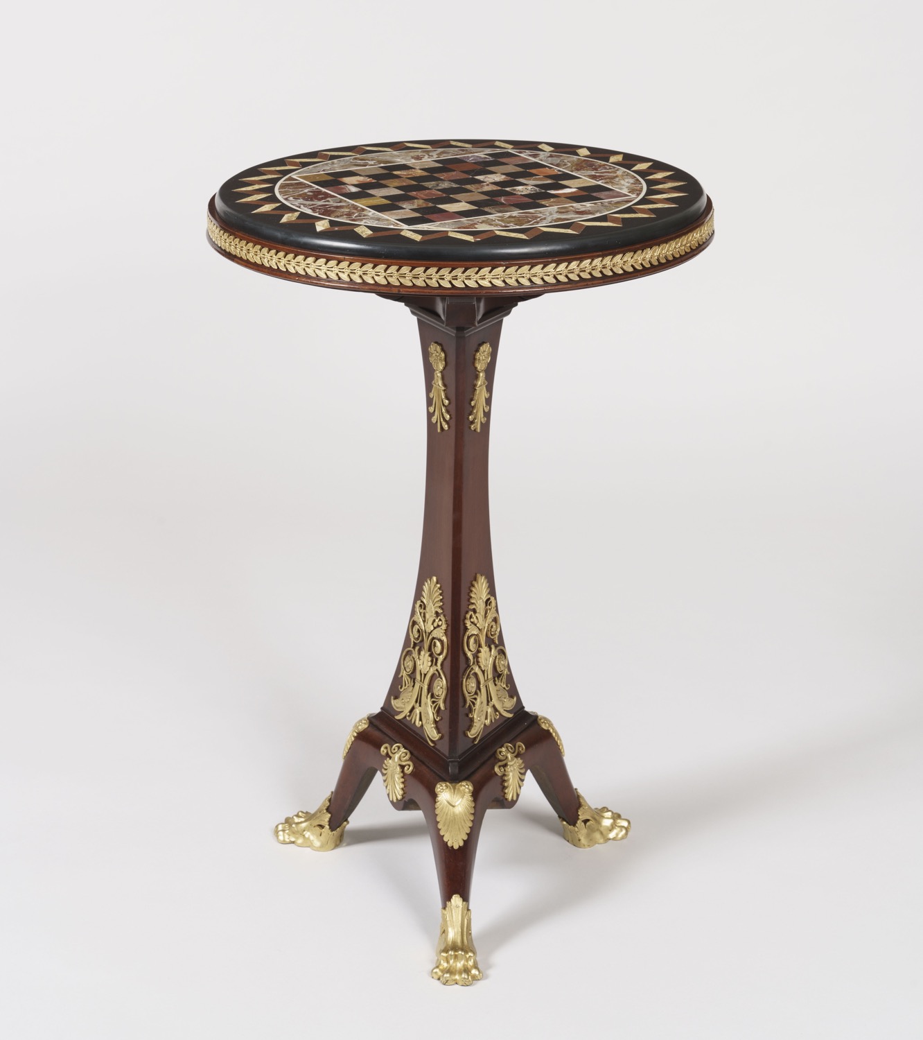 A high-quality specimen marble chess table (guéridon) featuring a circular top with a checkered game board made of various multicolored marbles. The mahogany pedestal base is richly decorated with gilded bronze (ormolu) floral mounts and terminates in a tripod base with gilded lion-paw feet.