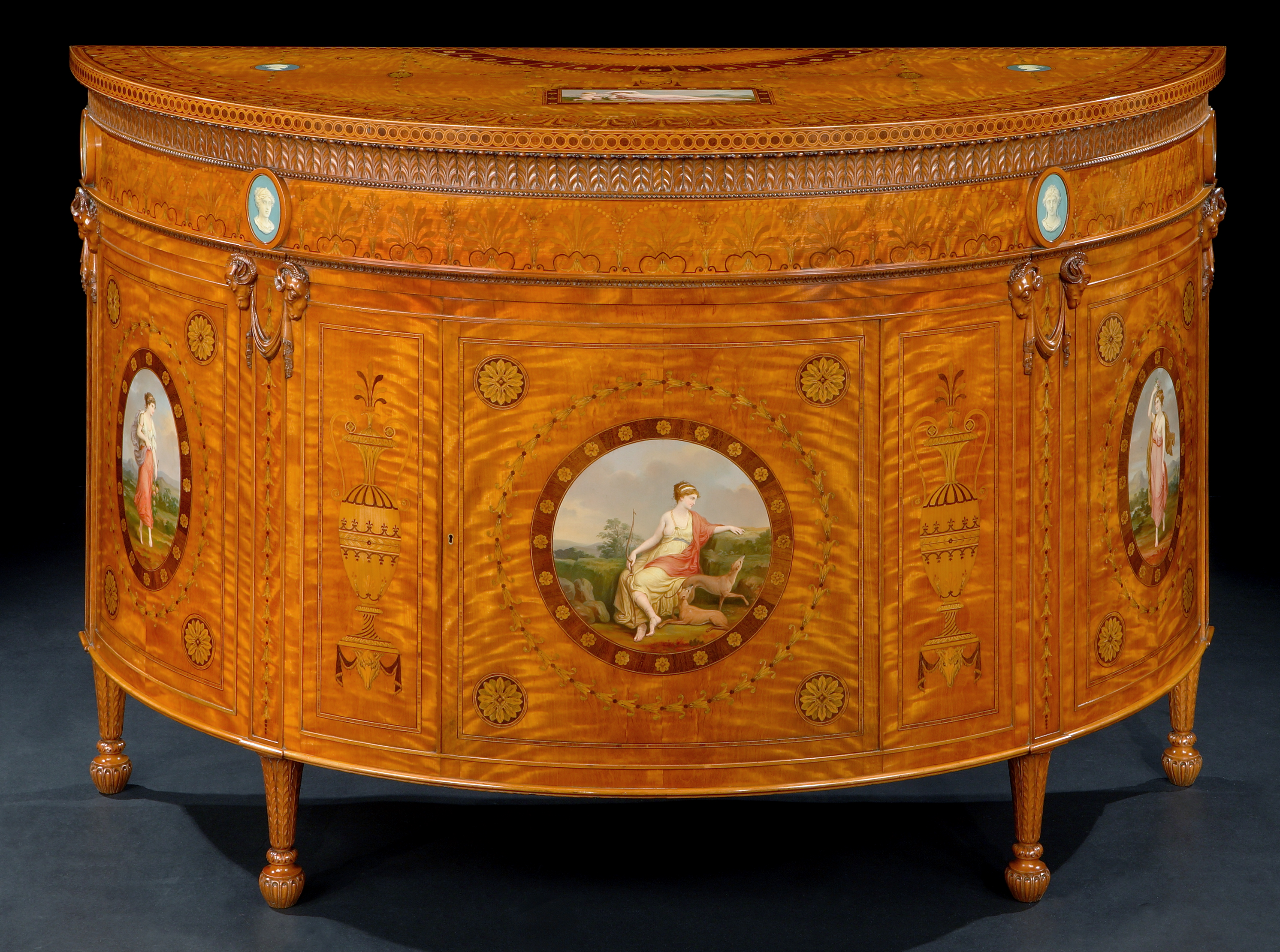 Semi-elliptical satinwood commode by Wright & Mansfield, decorated with neoclassical painted medallions of figures and urns. Features fine marquetry, Wedgwood-style blue jasperware plaques, and carved ram’s head details on tapered legs.