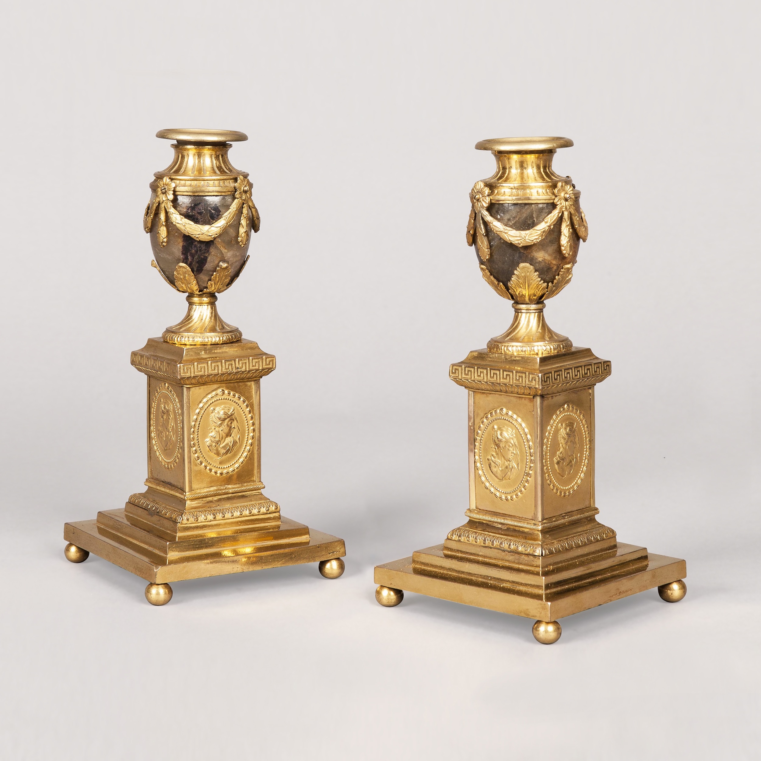 Two identical golden candle vases stand atop square pedestals with relief medallions and ball feet, featuring dark stone bodies draped in gilded swags against a seamless, light grey background.