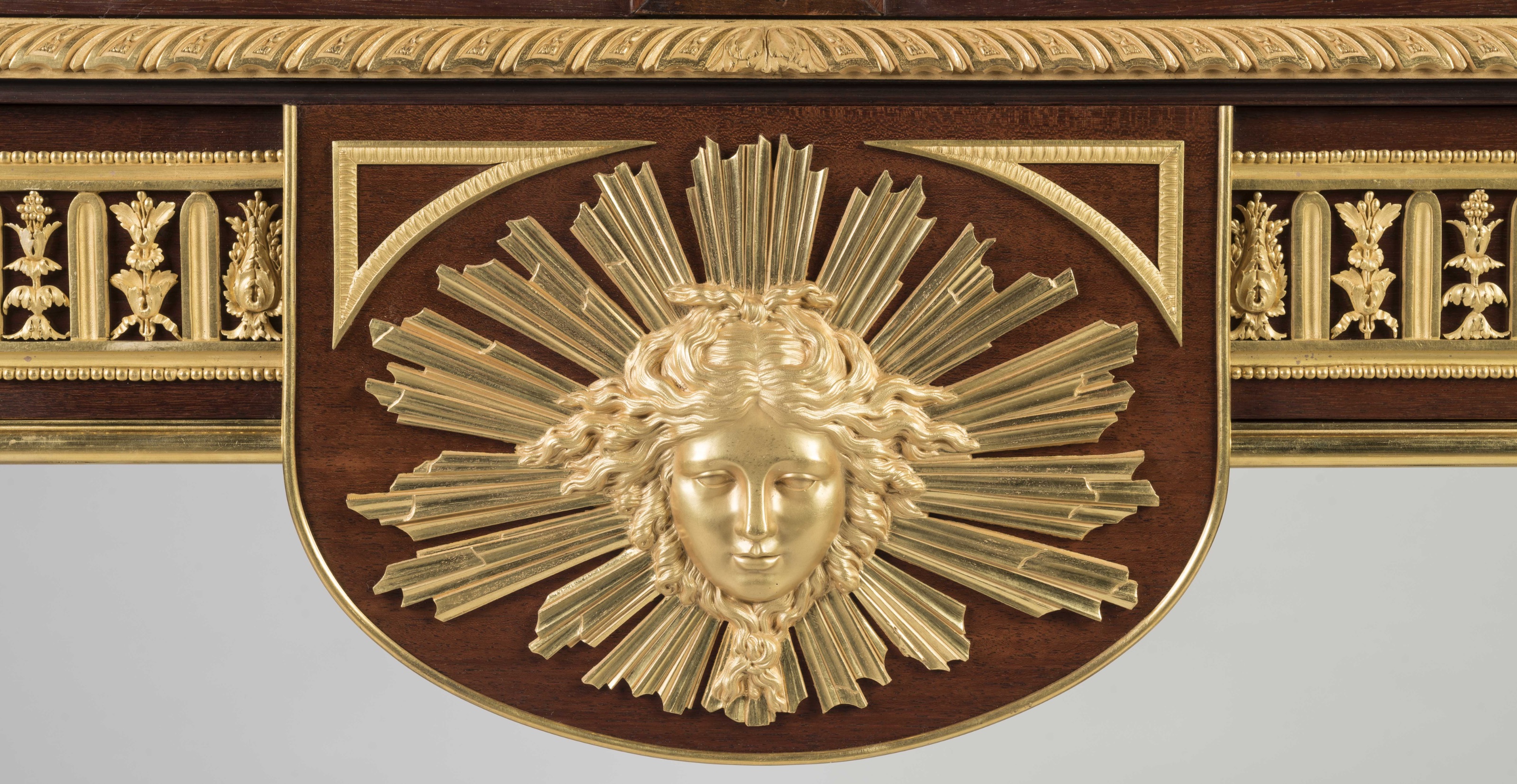A central gilded bronze sunburst mask with a serene face and radiating beams is mounted on a mahogany panel, flanked by ornate gold foliate borders and a decorative carved cornice.
