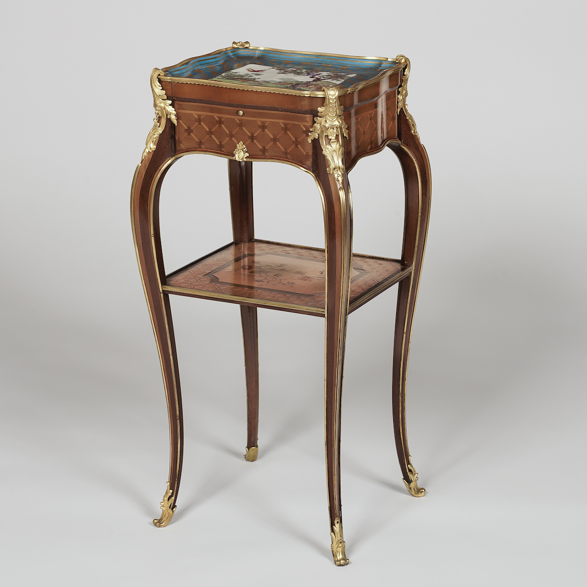 A slender marquetry occasional table with cabriole legs and a porcelain-inlaid top stands upright, featuring ornate gilt-bronze mounts and an undertier against a plain, neutral-toned background.