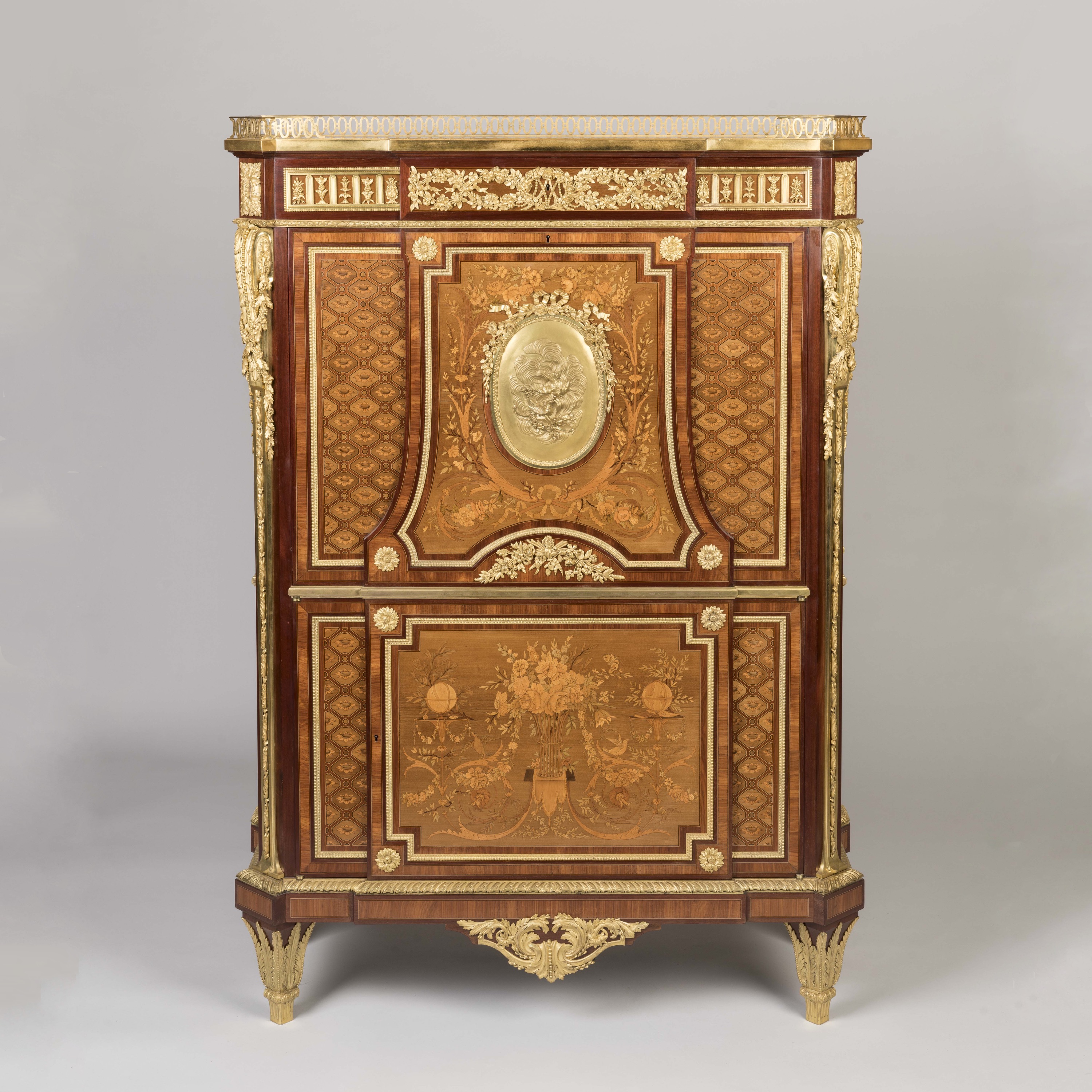 An ornate French Louis XVI style marquetry secrétaire à abattant (drop-front desk) crafted from fine wood with elaborate floral marquetry, gilt-bronze mounts, and a central medallion, set against a neutral grey background.
