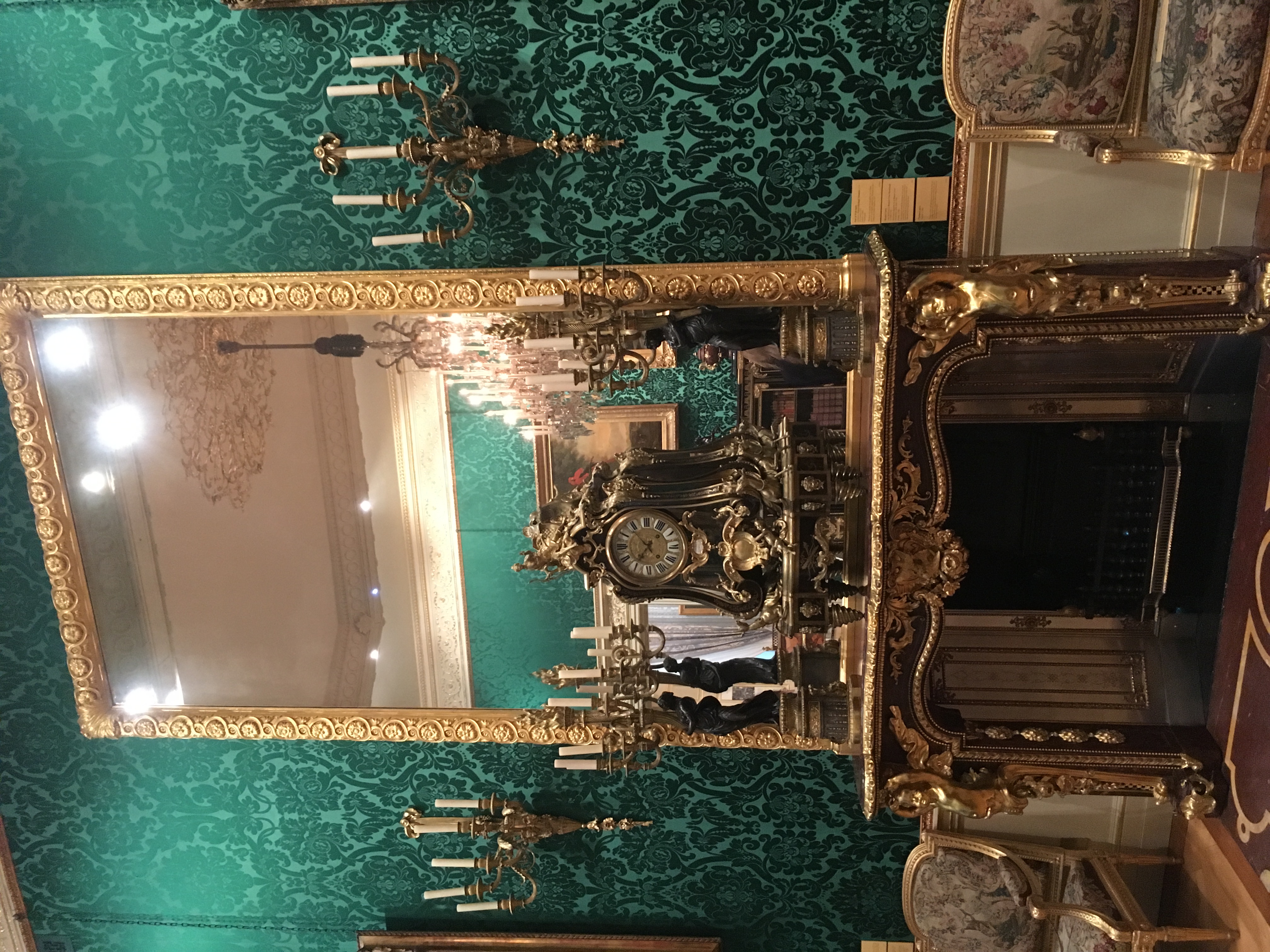 An ornate 18th-century style room interior featuring an elaborate gilt-wood mantelpiece, an oversized gilded mirror, an ornate mantel clock flanked by candelabras, and rich green damask wallpaper at the Wallace Collection.