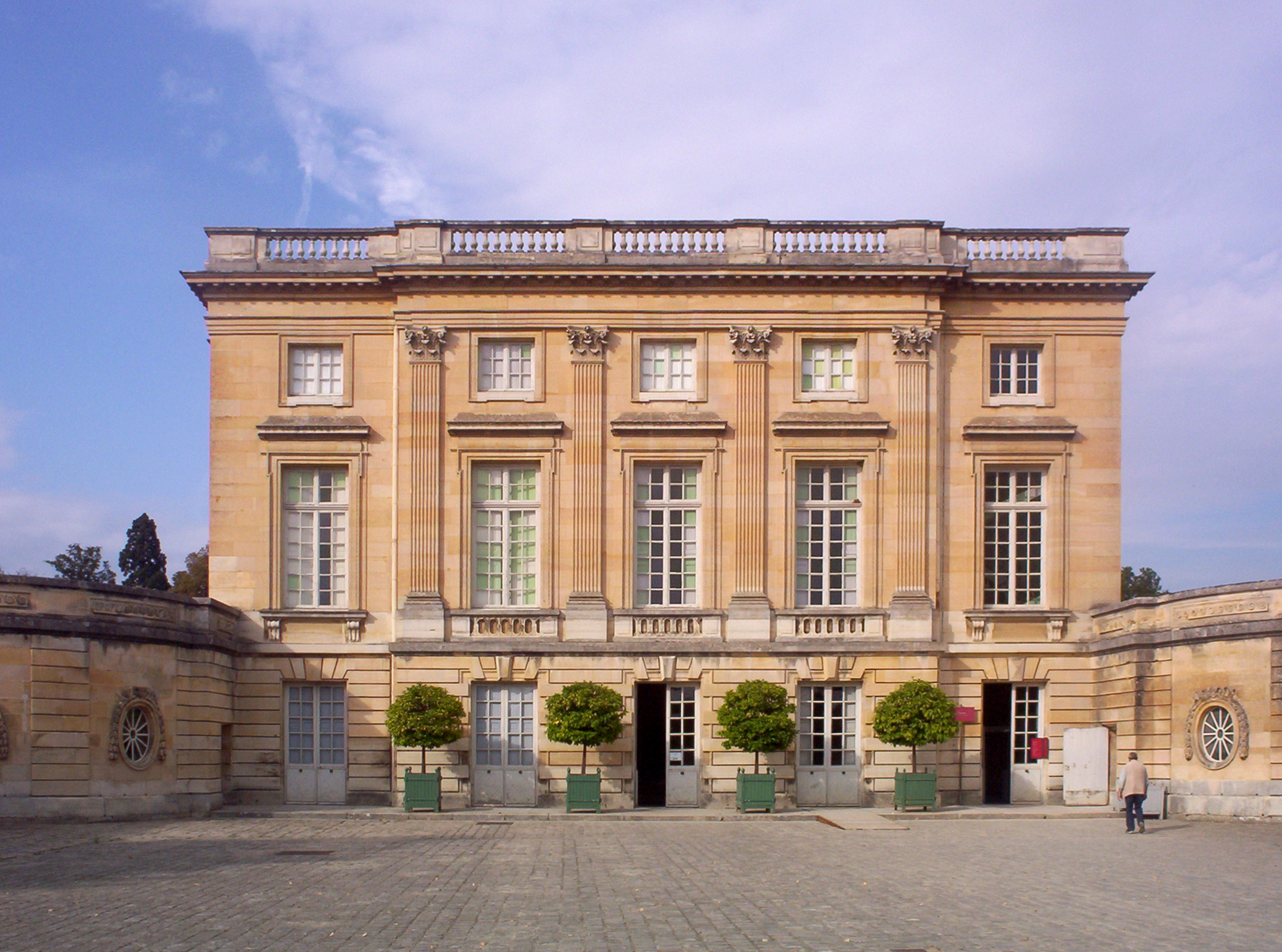 The grand, symmetrical light-yellow limestone facade of the Petit Trianon chateau at Versailles, framed by four Corinthian pilasters, set before a large paved cobblestone courtyard under a partly cloudy sky.