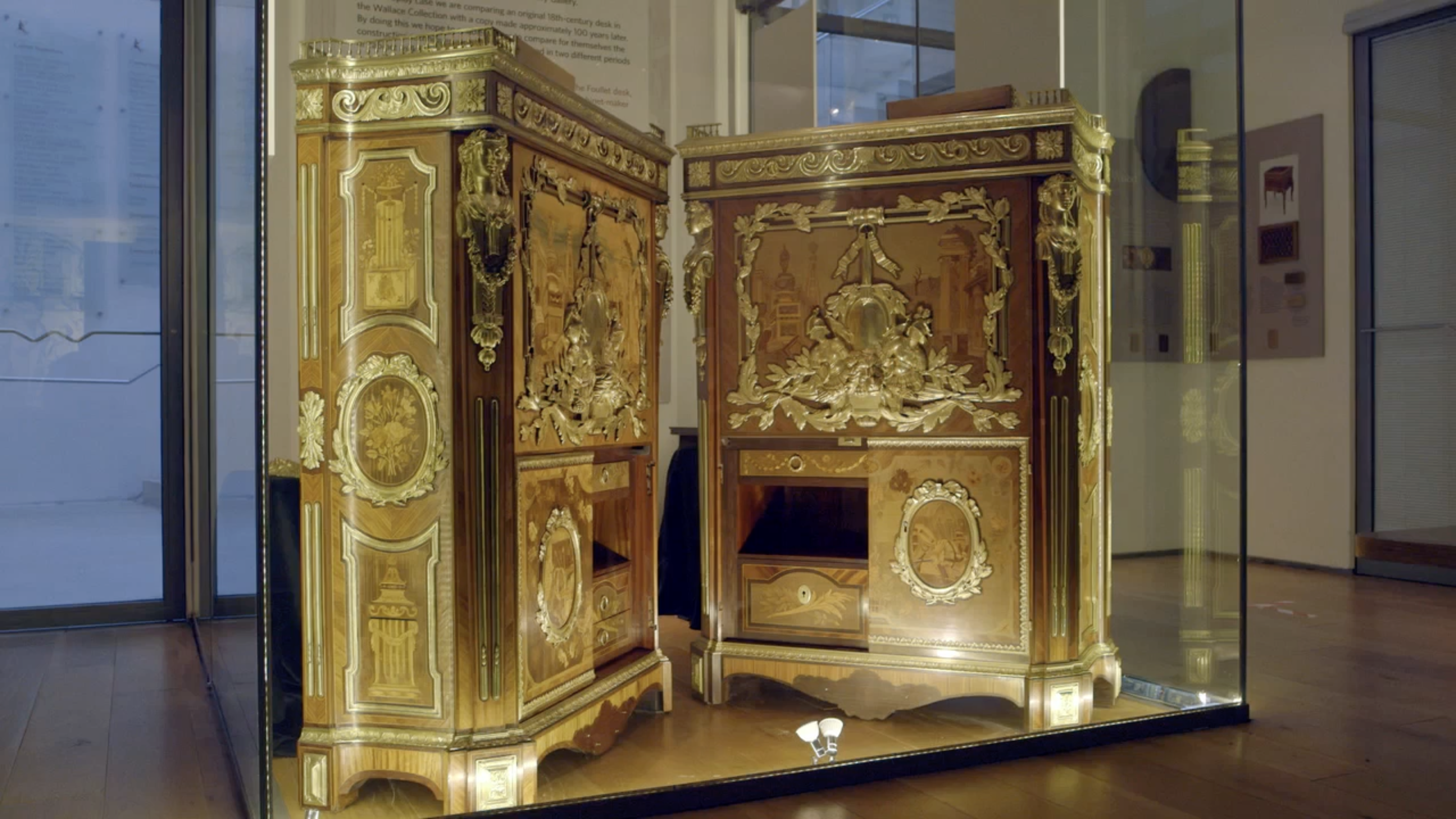 A side-by-side display of two highly ornate French 18th-century style marquetry secrétaires à abattant (drop-front desks) inside a glass museum exhibition case, showcasing elaborate wood marquetry, gilt-bronze mounts, and central medallions.