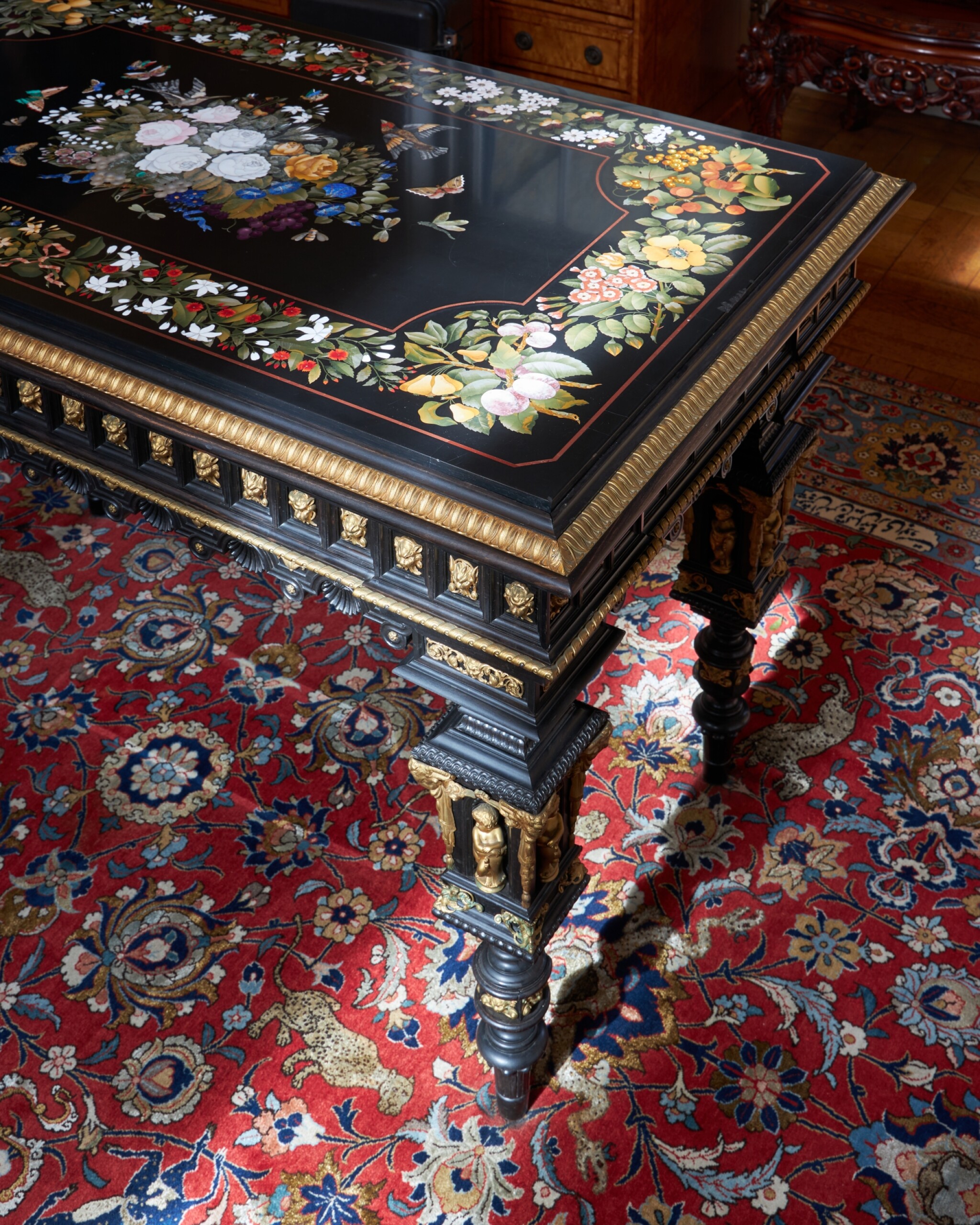 Ornate Pietra Dura and ebonized wood library table. The black marble top features intricate floral and bird inlays, supported by a gilt-mounted base with carved figures over a red patterned rug.